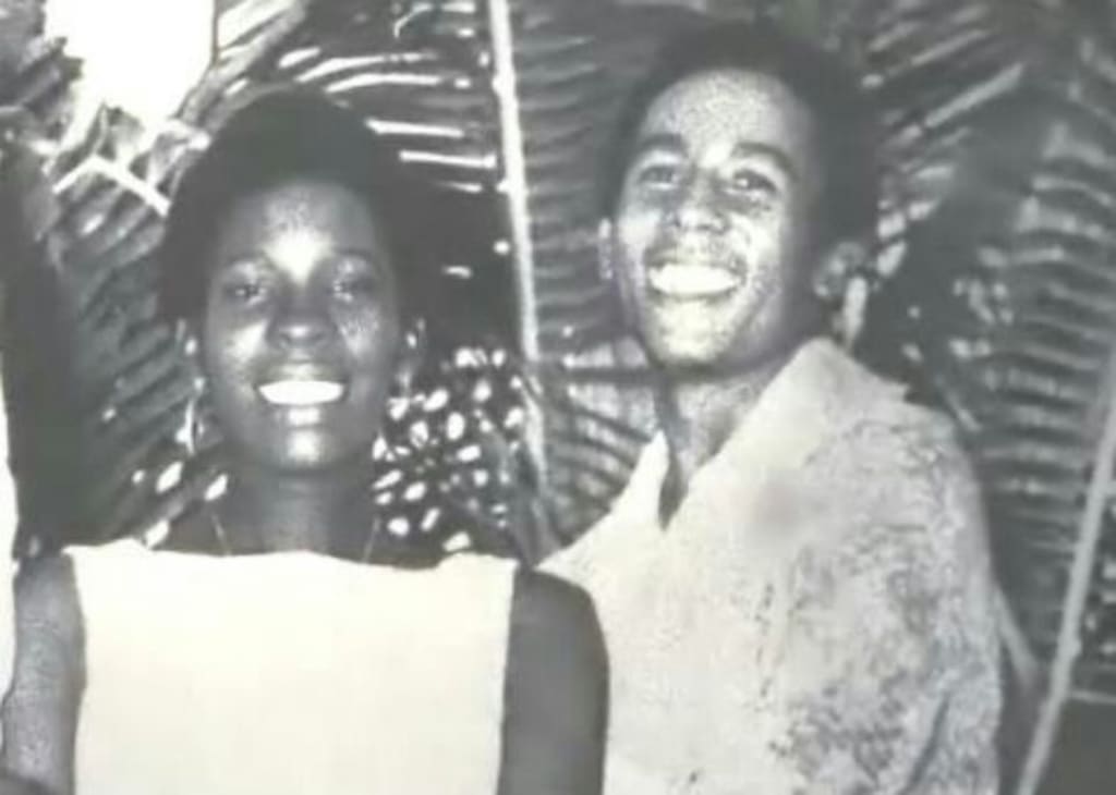 A young Bob Marley and Rita Marley in a black and white photo standing under a palm tree smiling