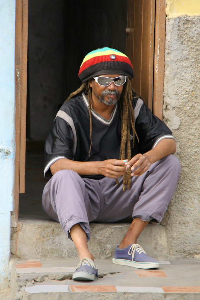 Rastafarian man with red, gold, and green tam hat on sits in a concrete doorway with dreadlocks and sunglasses